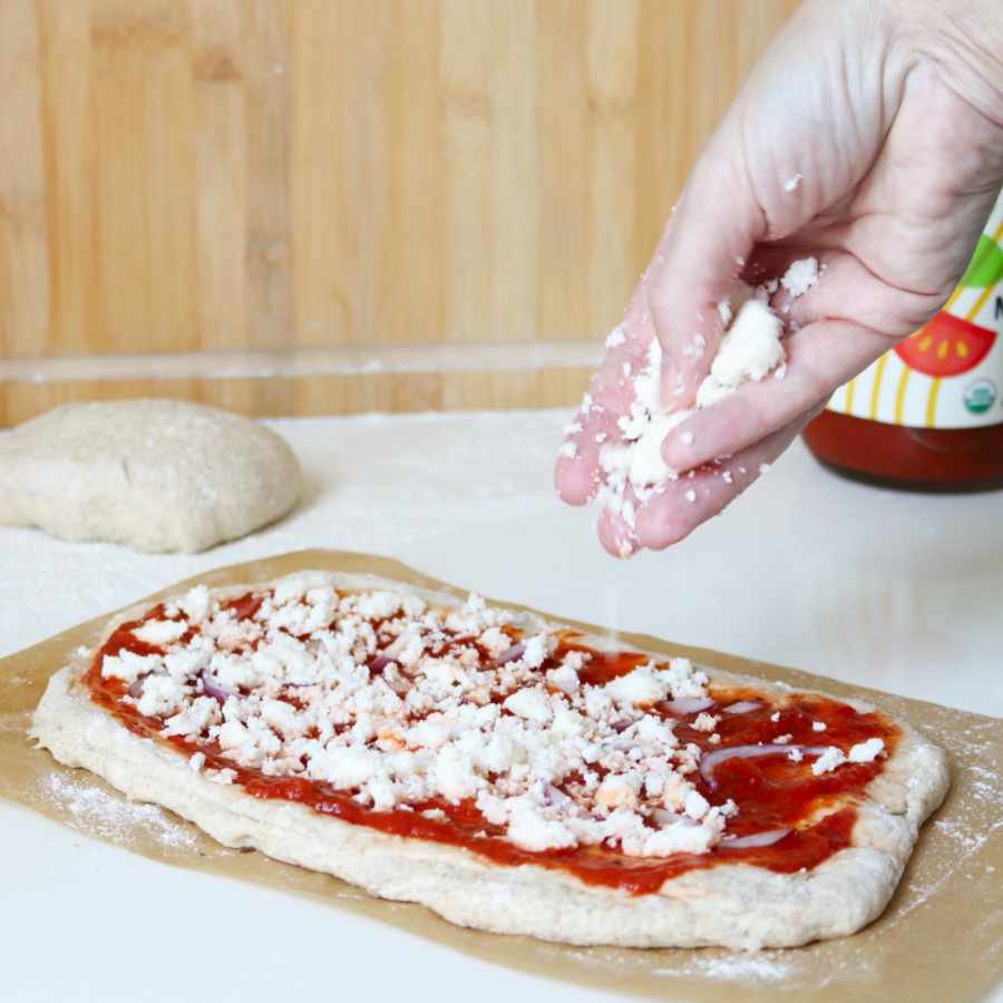 hand sprinkles white cheese crumbs on a raw shaped pizza dough with tomatoe sauce - Breadista