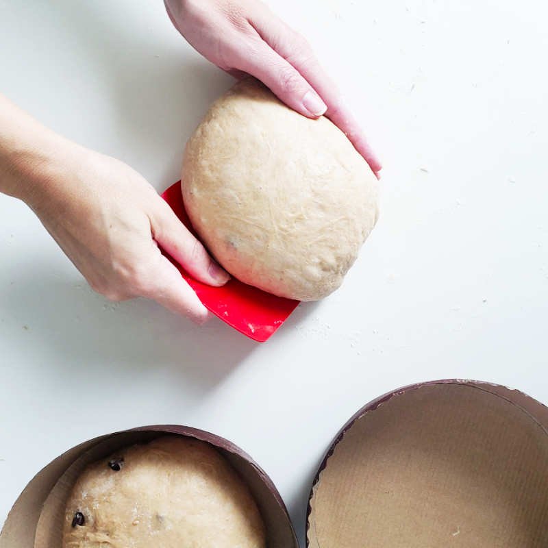 Birdview - two hands shaping floured dough ball round while one hand holds red dough scraper by Breadista