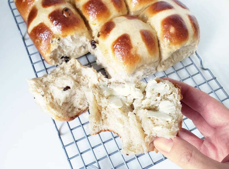 primary view - Two halves of hot cross buns, one with butter is hold by right hand
