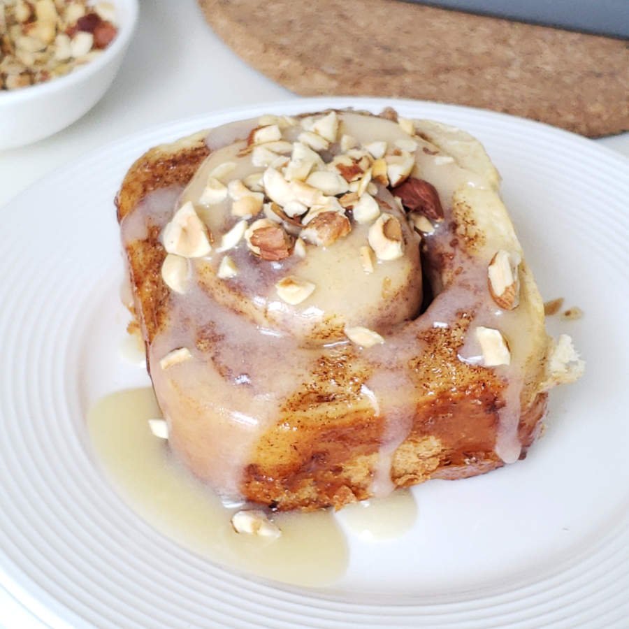 Homemade fluffy Cinnamon Roll on plate drizzled with light frosting and topped with chopped hazelnuts