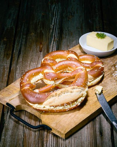 2 Butterbrezel on wooden board with knife and piece of butter on small plate by Schaefer dein Baecker