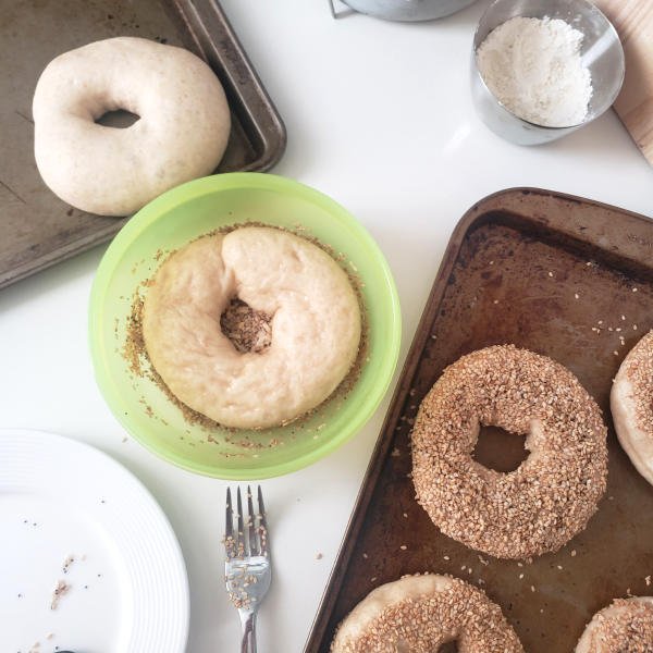 Bagels getting topping - some already topped, one upside down in container with seeds, another raw & risen on another baking sheet - by BREADISTA made with a bread mix