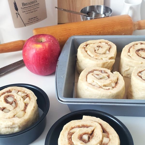 closeup - big cut apple-cinnamon rolls unbaked in square pan and two single pans. Next to red apple and wooden rolling pin - by BREADISTA