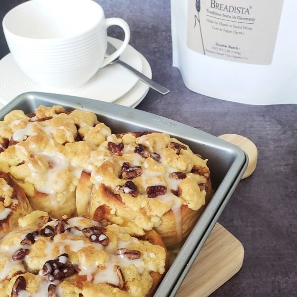 closeup - big cinnamon rolls baked with streusel-nut topping in pan, white coffee cup on plate in the back - by BREADISTA