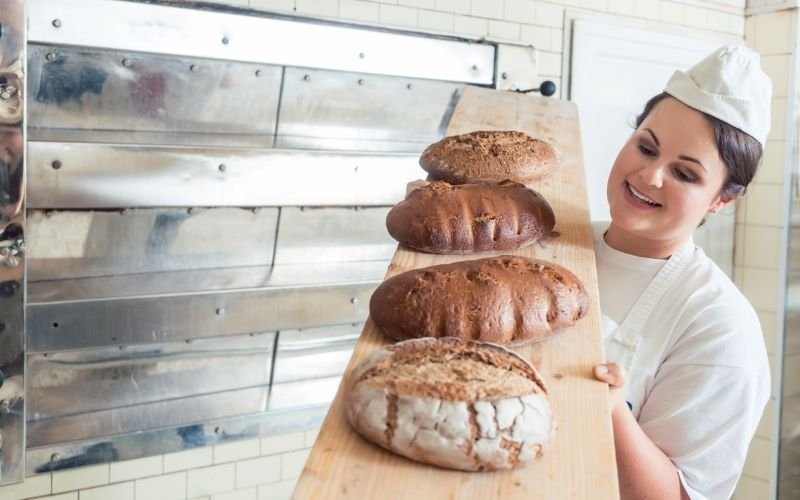 Female Baker with 4 different loaves on panel in front of commercial oven. German bread culture - BREADISTA